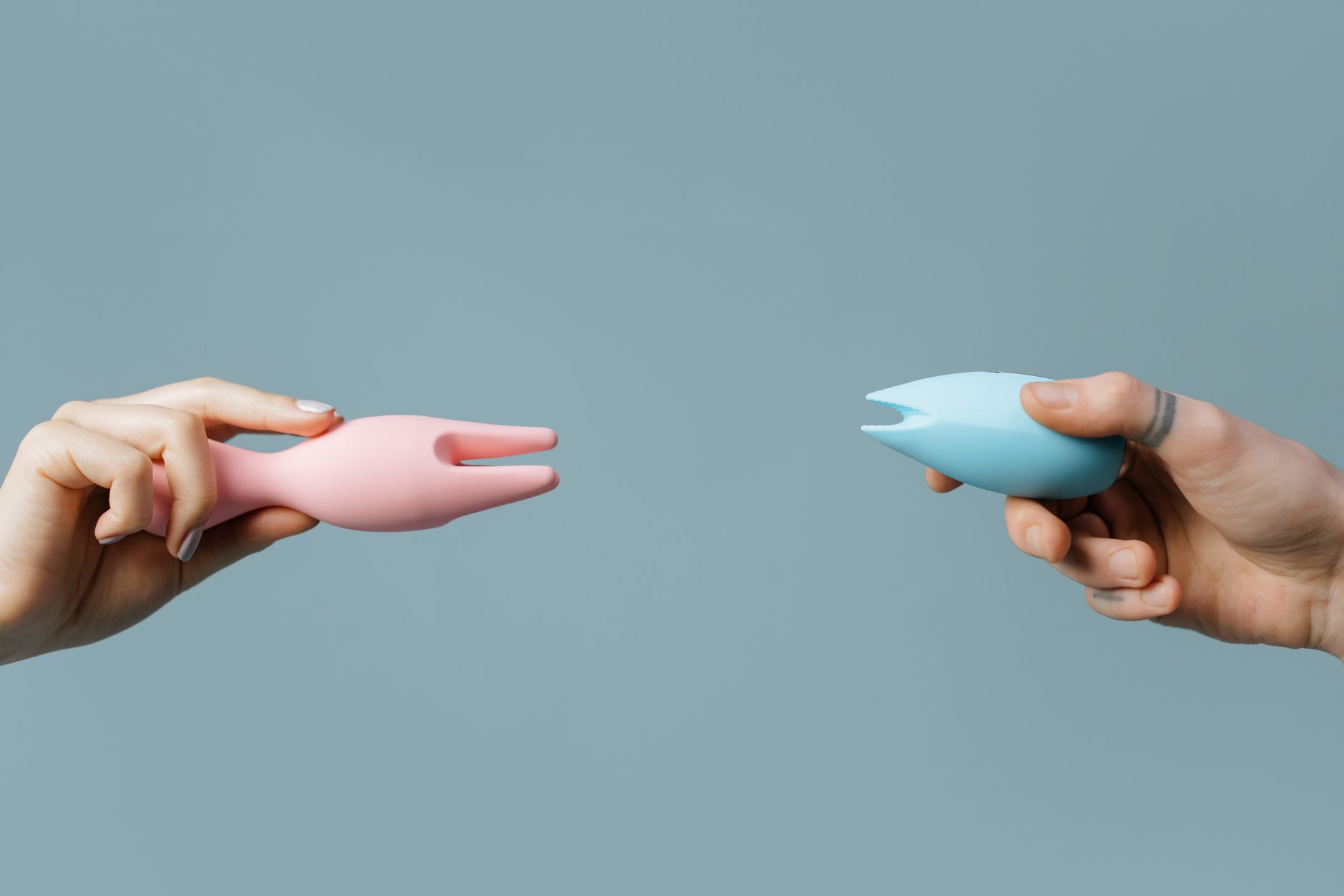 Two hands holding pink and blue silicone vibrators against a pastel background