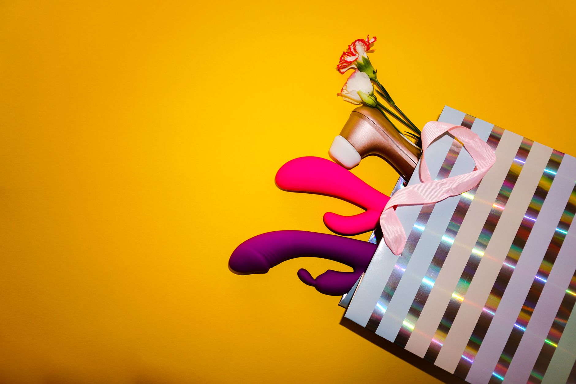 Colorful vibrators with flowers in a holographic gift bag on a bold yellow background.